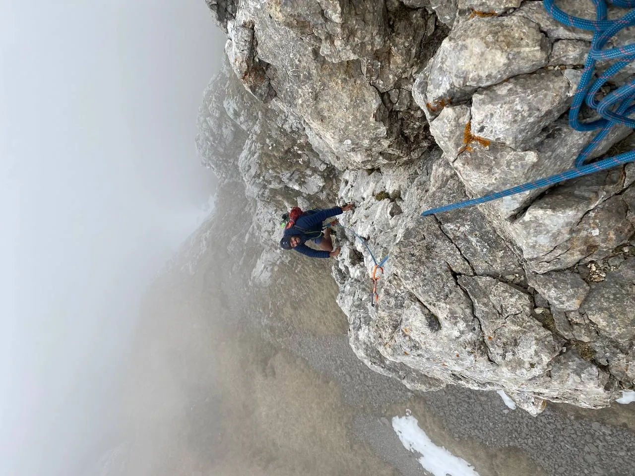 Looking steeply down the grey limestone of a Galtigen tower as a climber leads on the blue rope, the wall dropping away into fog below.