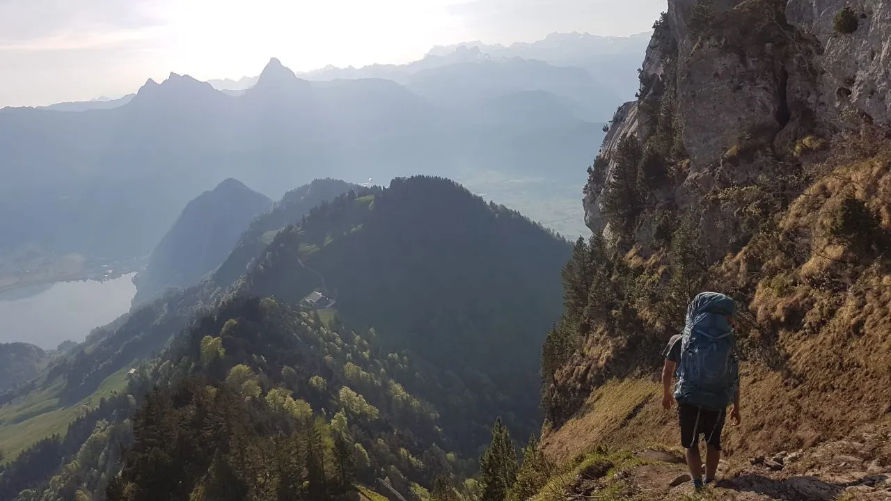 A hiker with a large pack walks an exposed grassy traverse below a rock wall, hazy ridges and a lake far below in the low sun.