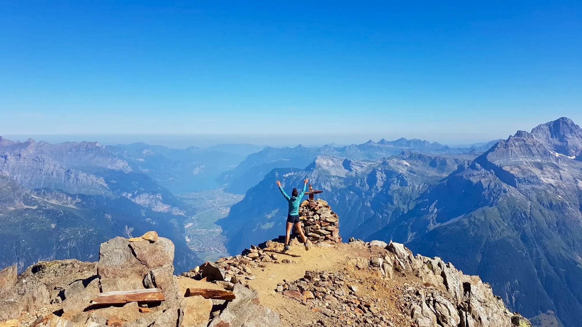 Una mujer en la cumbre del Bristen con los brazos levantados junto al hito de piedras, mirando los Alpes de Uri y de Glaris.