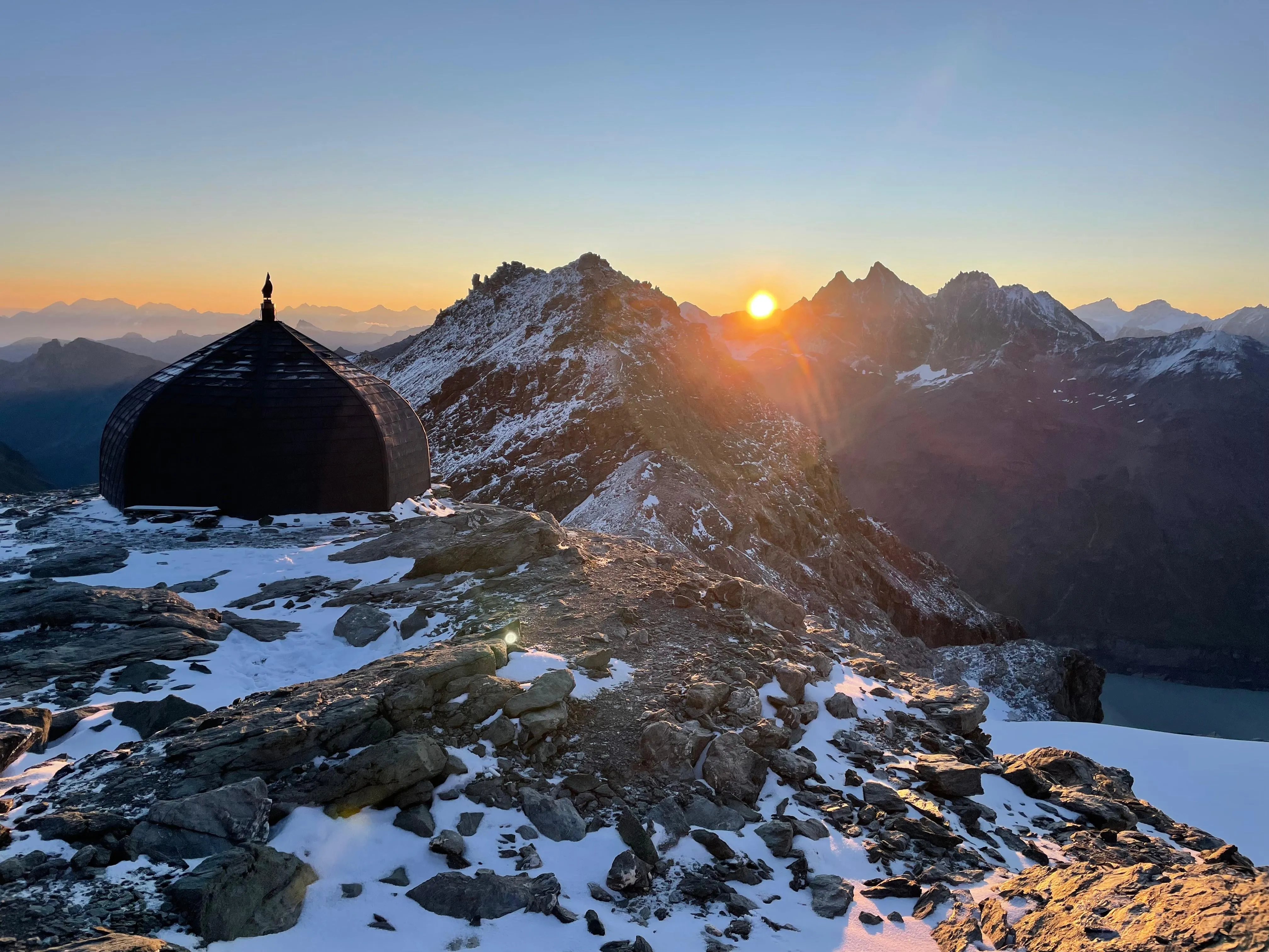Una cabaña negra con forma de cúpula en una cresta nevada mientras el sol sale sobre cumbres lejanas.