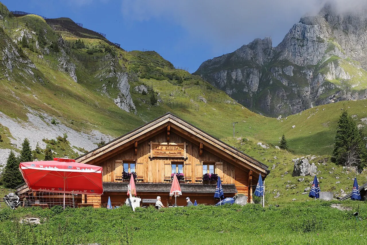 The Alp Rigidal chalet on a green alpine meadow above Engelberg, with steep, rugged limestone cliffs behind under a blue sky.