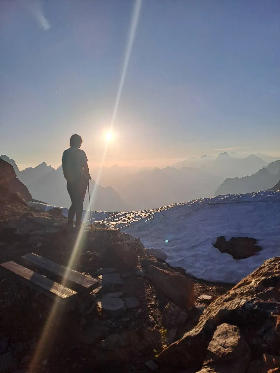 A hiker stands silhouetted on a rocky ledge looking into the sunrise over hazy mountain ranges, a snowfield beside her.
