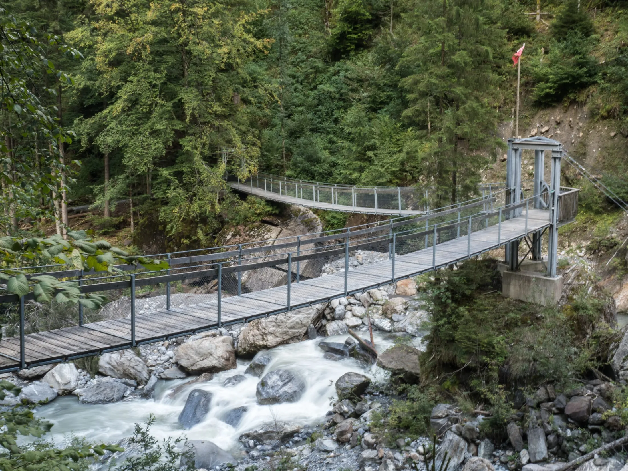 A wooden suspension footbridge crossing the wild Engelberger Aa in the gorge, with a second bridge beyond and whitewater tumbling over boulders below.