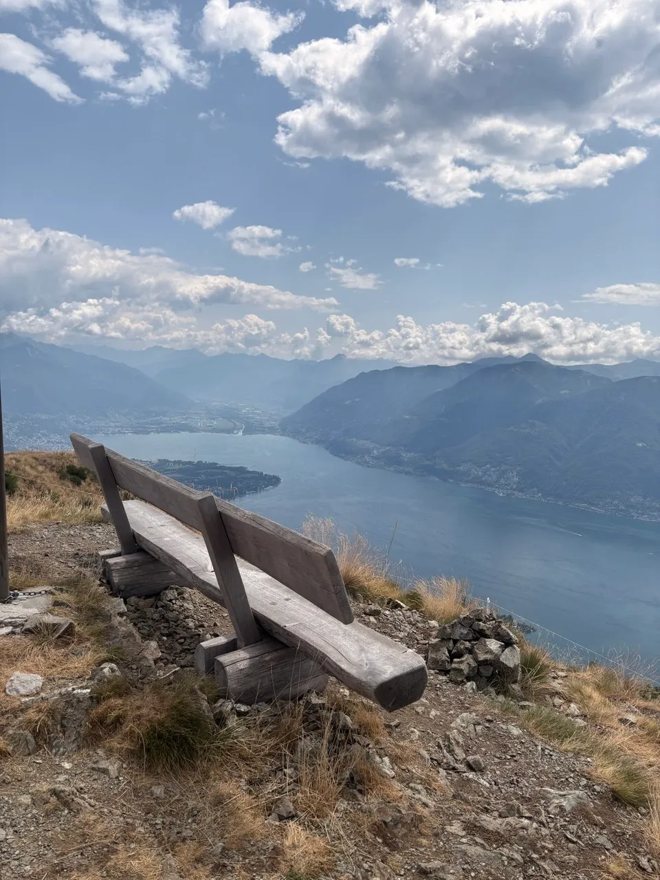 A weathered wooden bench on a grassy viewpoint looking out over Lago Maggiore and the Locarno delta, hazy mountains all around.