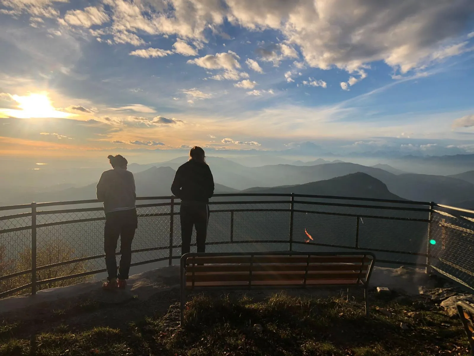 Two people stand at a viewpoint railing gazing across hazy mountain ranges toward a low sun, with a bench in the foreground.