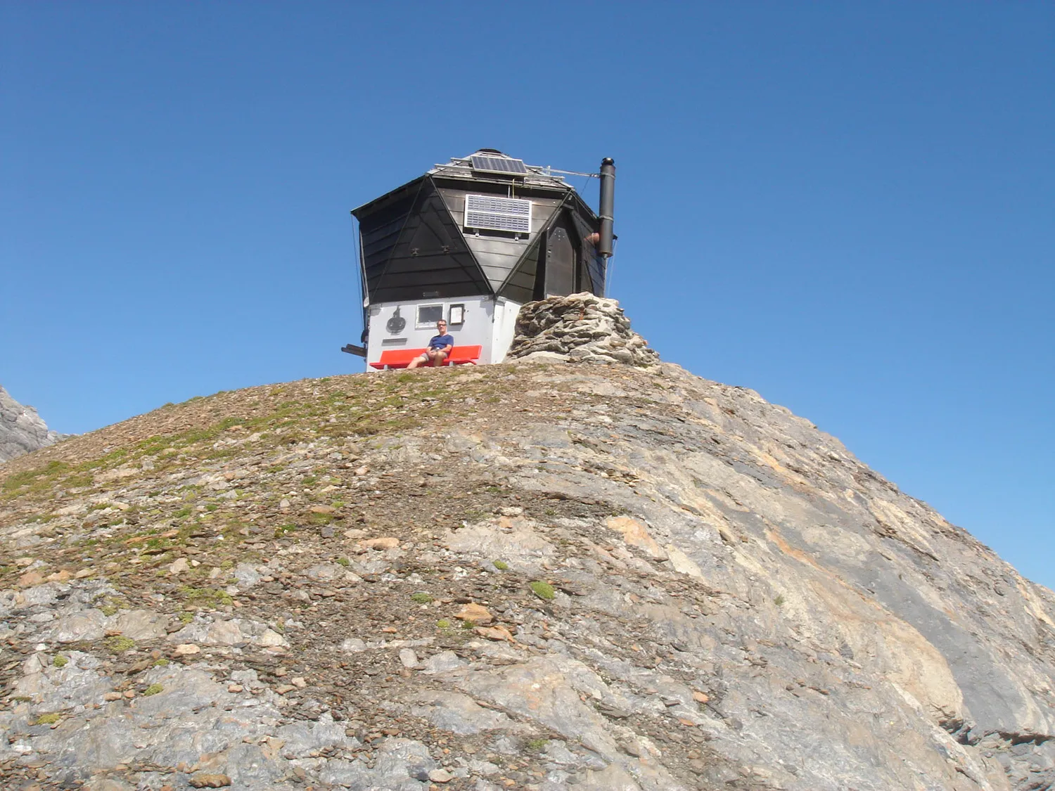 The small Grassen bivouac perched on a rocky knoll under a blue sky, with a person sitting out front in the sun.