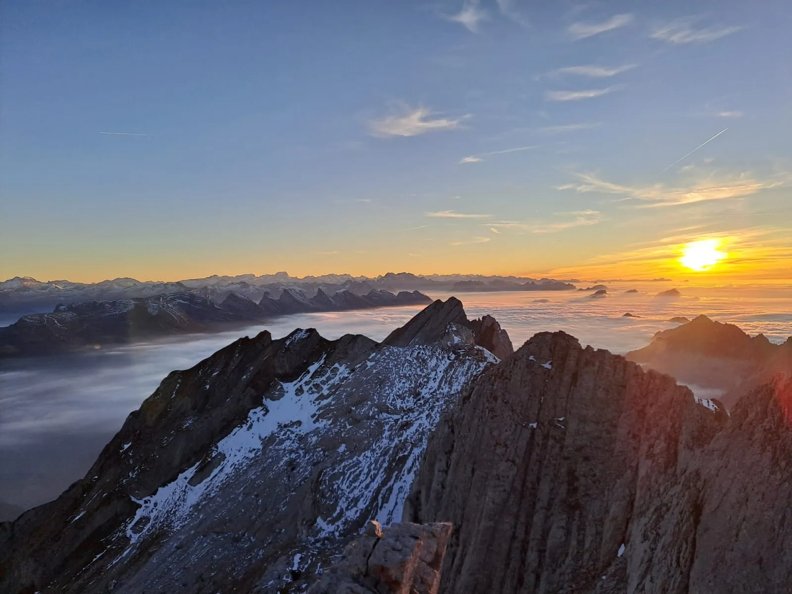 An autumn sunset over a sea of fog, the Alpstein ridge in the foreground and the Alps on the horizon.