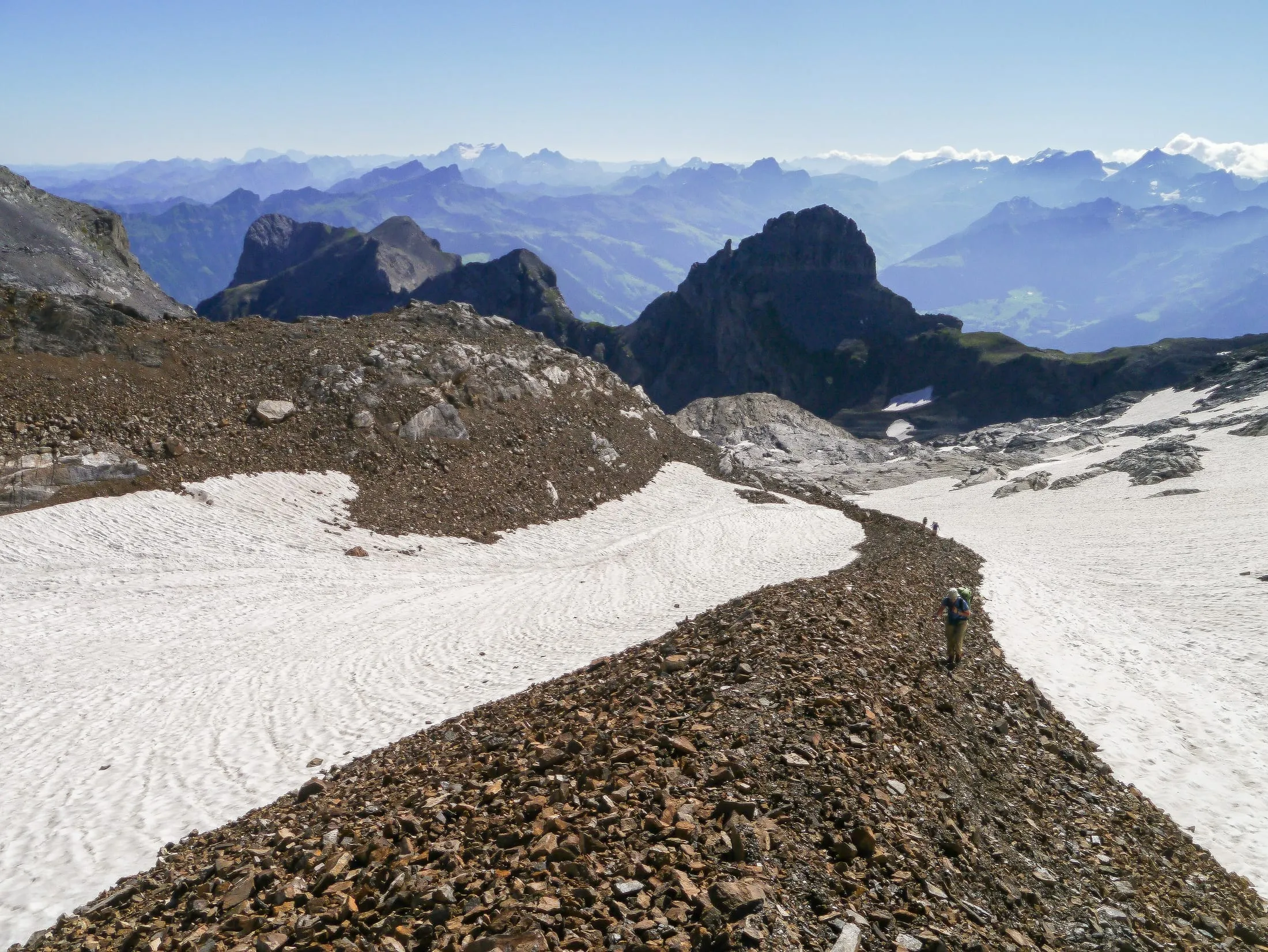 A hiker on the scree crest between the firn fields on the Uri Rotstock, with the Central Swiss pre-Alps beyond.