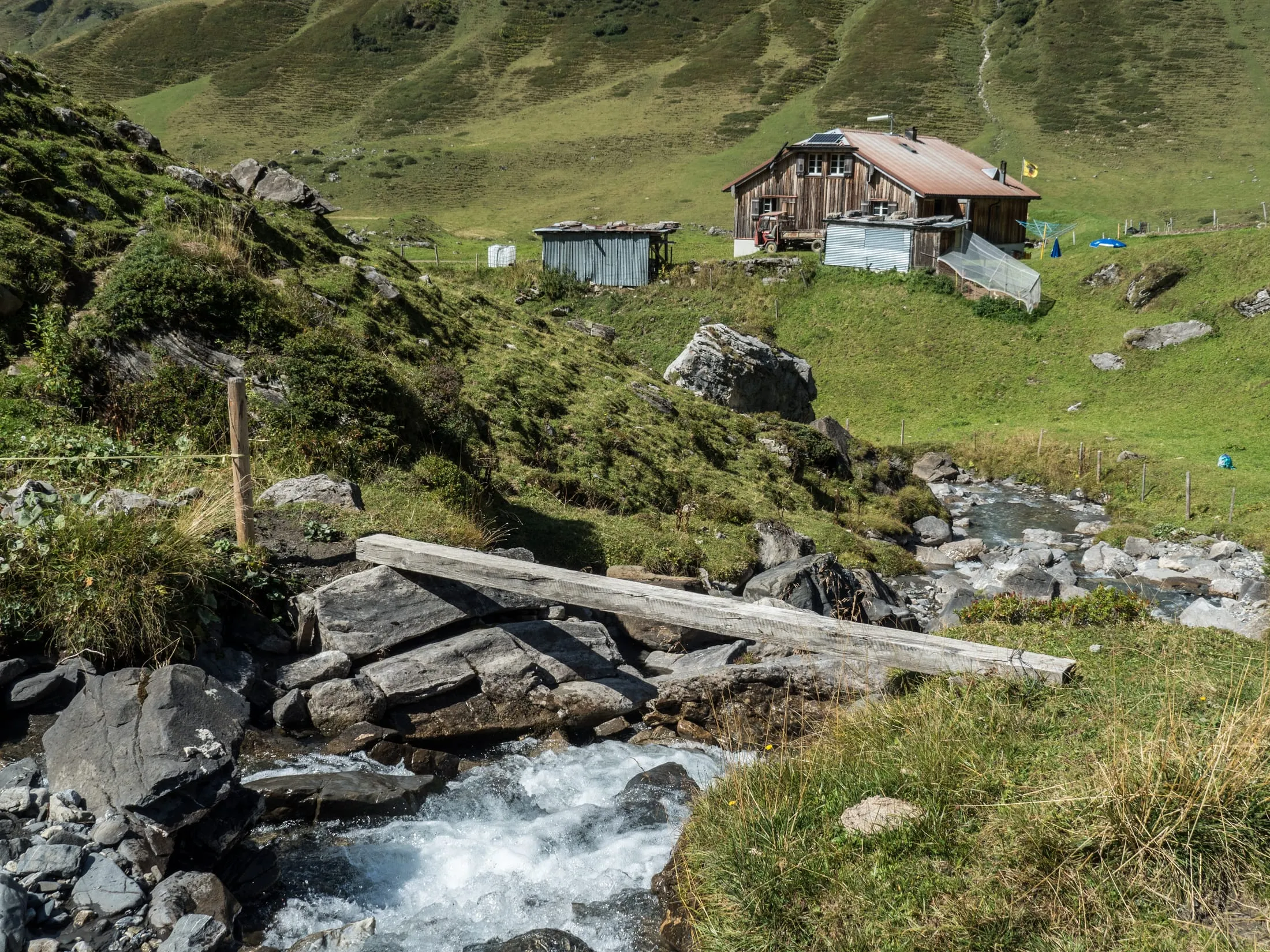 A wooden plank footbridge over the rushing Stierenbach stream, with the Blackenalp alp building on green pasture behind, framed by rocky mountain slopes.