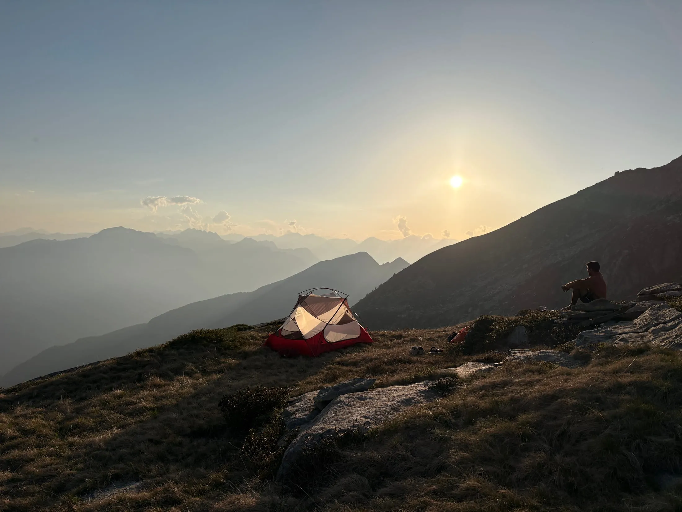 A small tent pitched on a grassy ridge at golden hour, a hiker sitting on a rock nearby as the sun sinks behind hazy Ticino peaks.