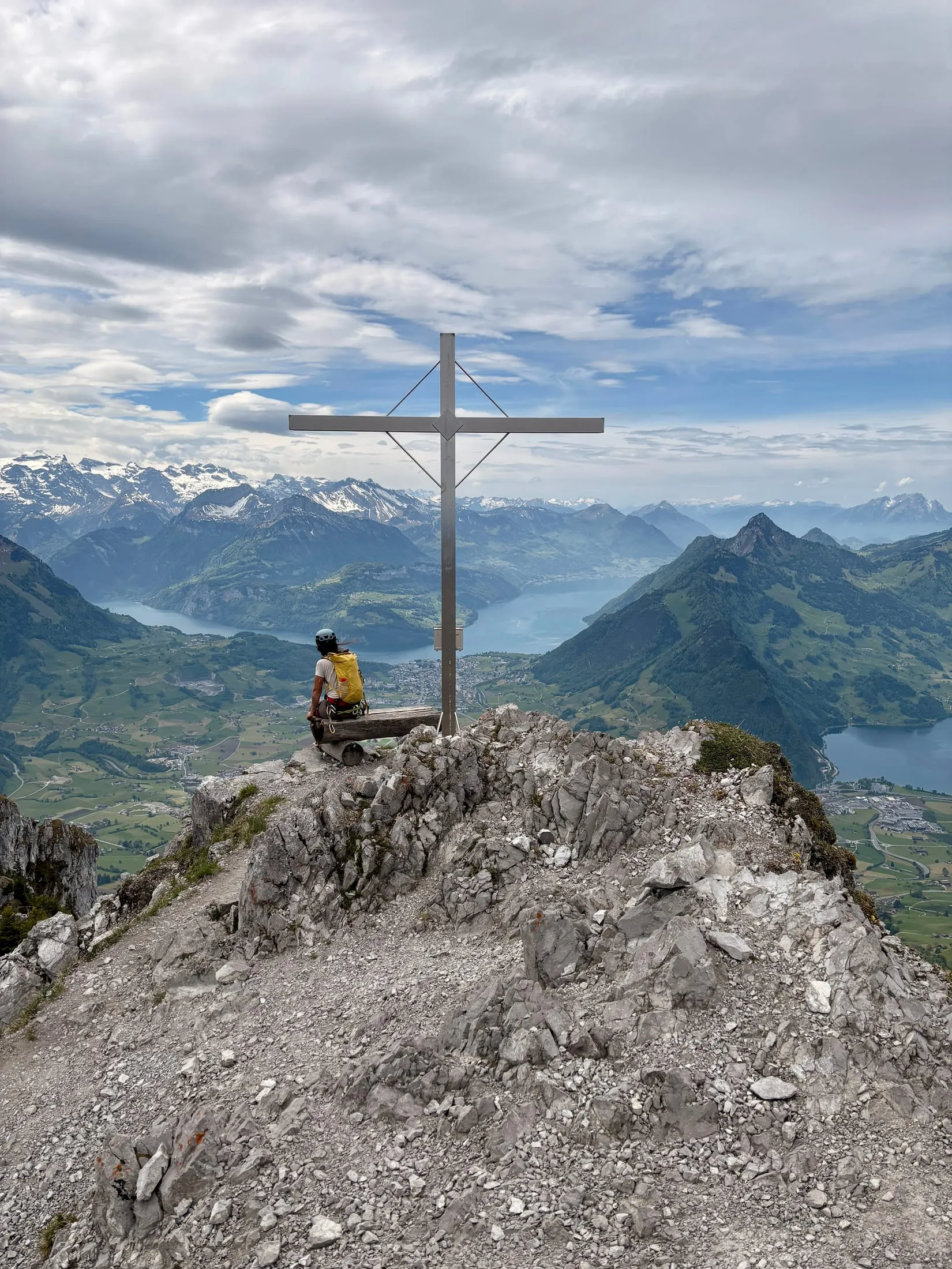 A hiker rests beside the summit cross of the Haggenspitz, with the snow-streaked Urner Alps and the Lake Lucerne basin spread out behind.