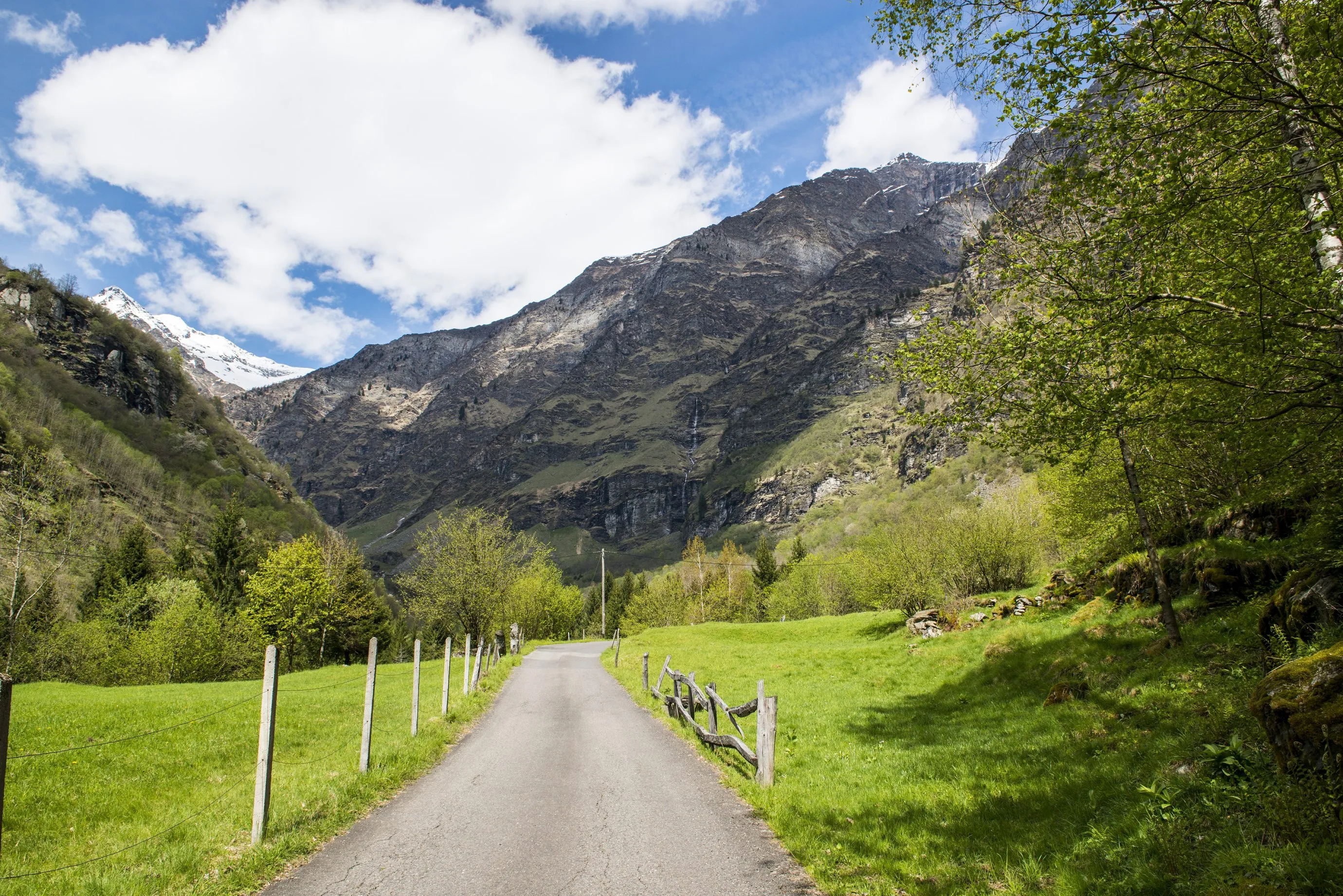 A valley road across green meadows toward the rock walls of the Cima Rossa massif, Val Malvaglia in Ticino, early summer.