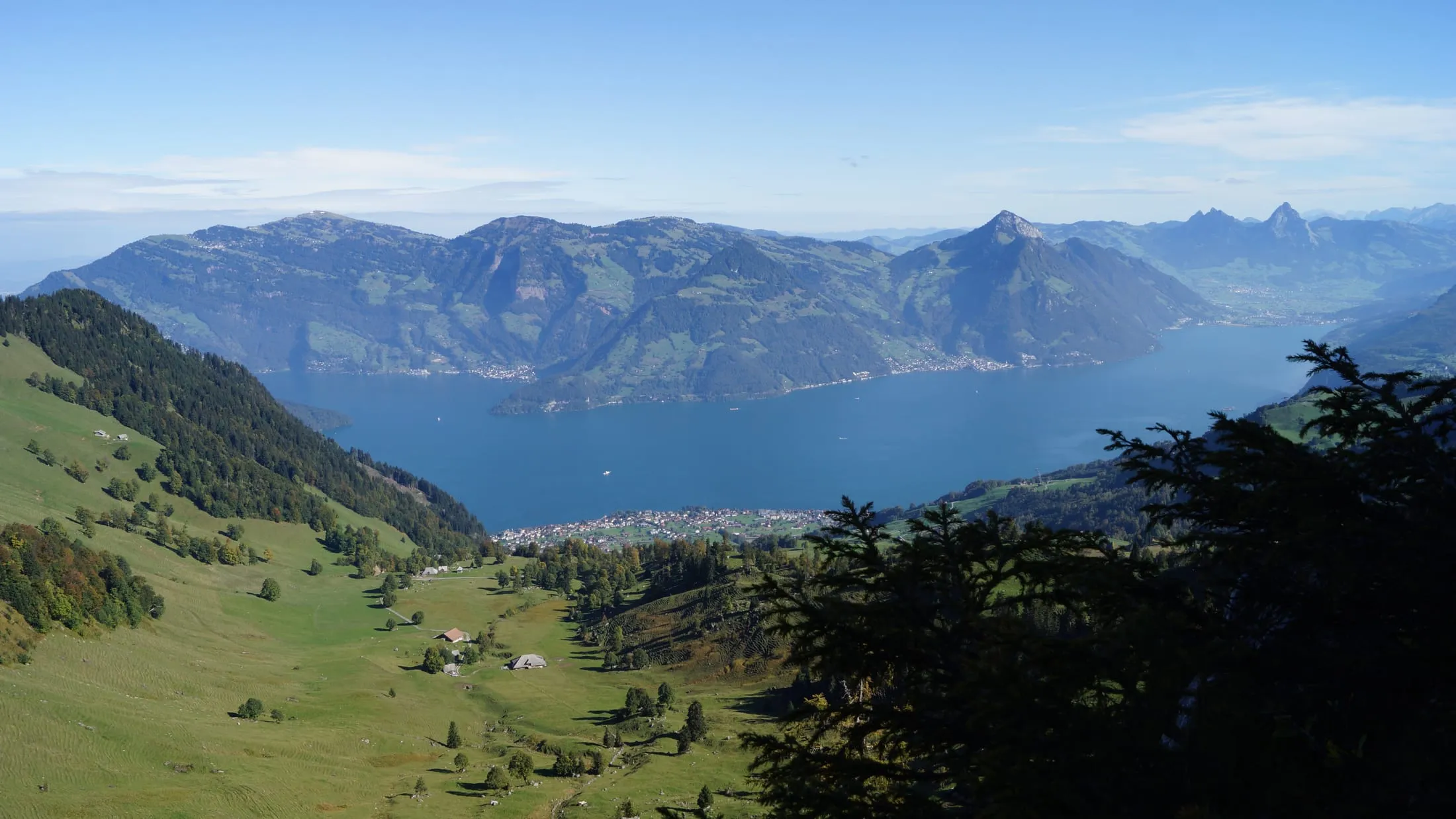 View from the Bleikigrat over green alpine meadows down to Beckenried on the deep-blue Lake Lucerne, with the Rigi behind and the Mythen peaks to the right.