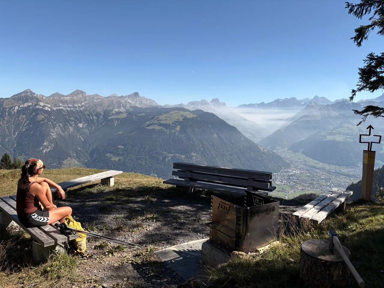A hiker sits on a wooden bench at the Wanggrat barbecue spot, looking out over the Reuss valley and the Uri Alps in the morning haze.