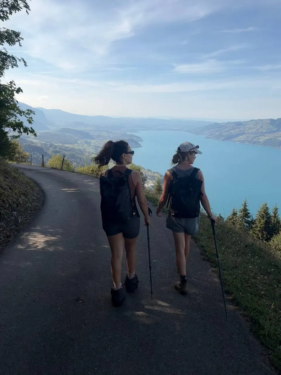 Dos excursionistas con bastones bajan por una carretera estrecha muy por encima del turquesa lago de Thun.