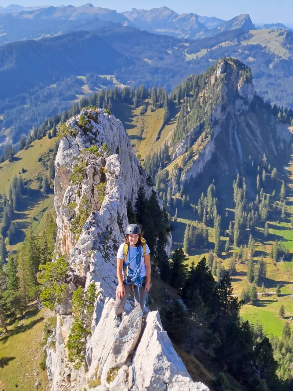 A helmeted climber in a T-shirt, rope racked, on the narrow Brotmessergrat, with the white rock blade and the green Prealp slopes of the Pilatus massif behind.