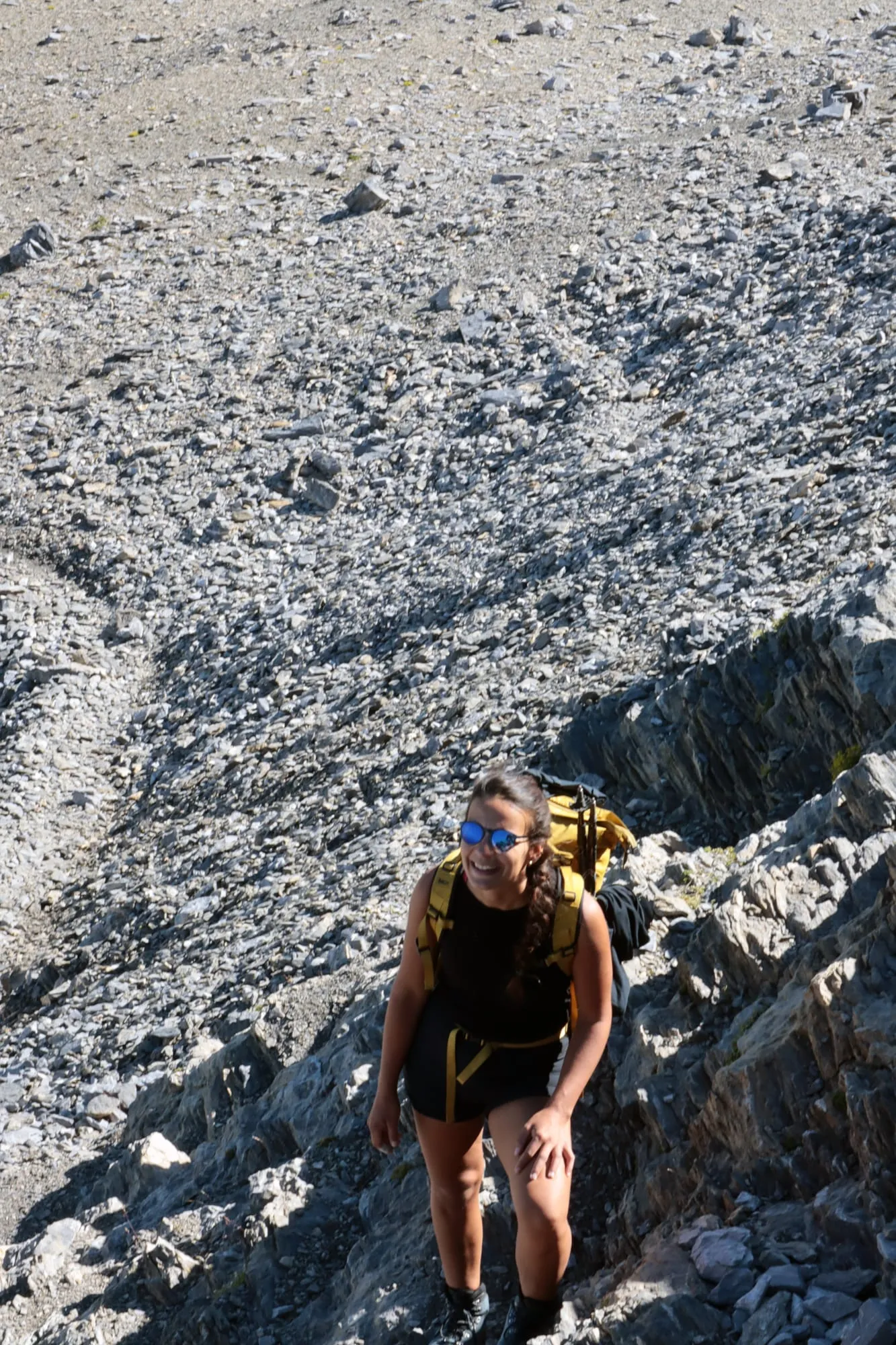 A mountaineer with a yellow pack and sunglasses laughs while climbing up the pale scree slope.
