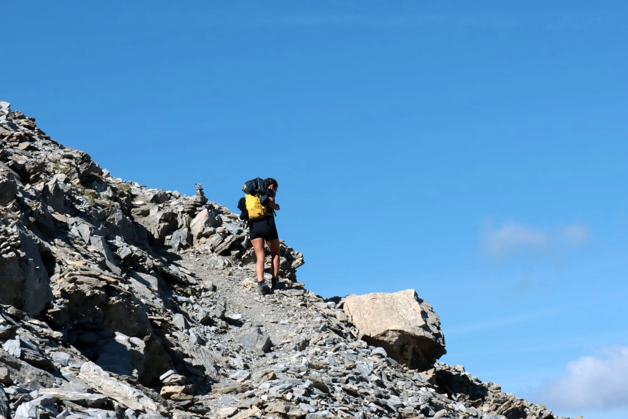 A mountaineer on the scree path to the ridge, nothing above her but blue sky and a cairn.
