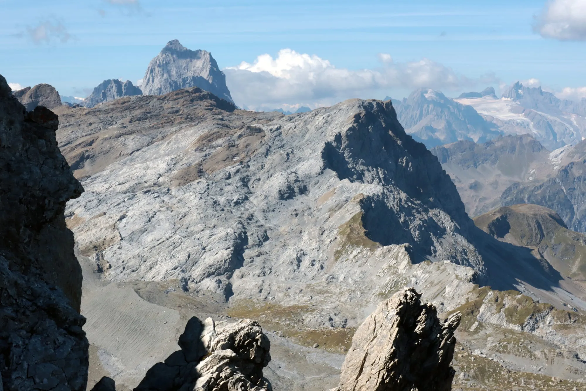 A view over pale limestone slabs to a sharp rock tooth, with distant glaciated peaks behind.