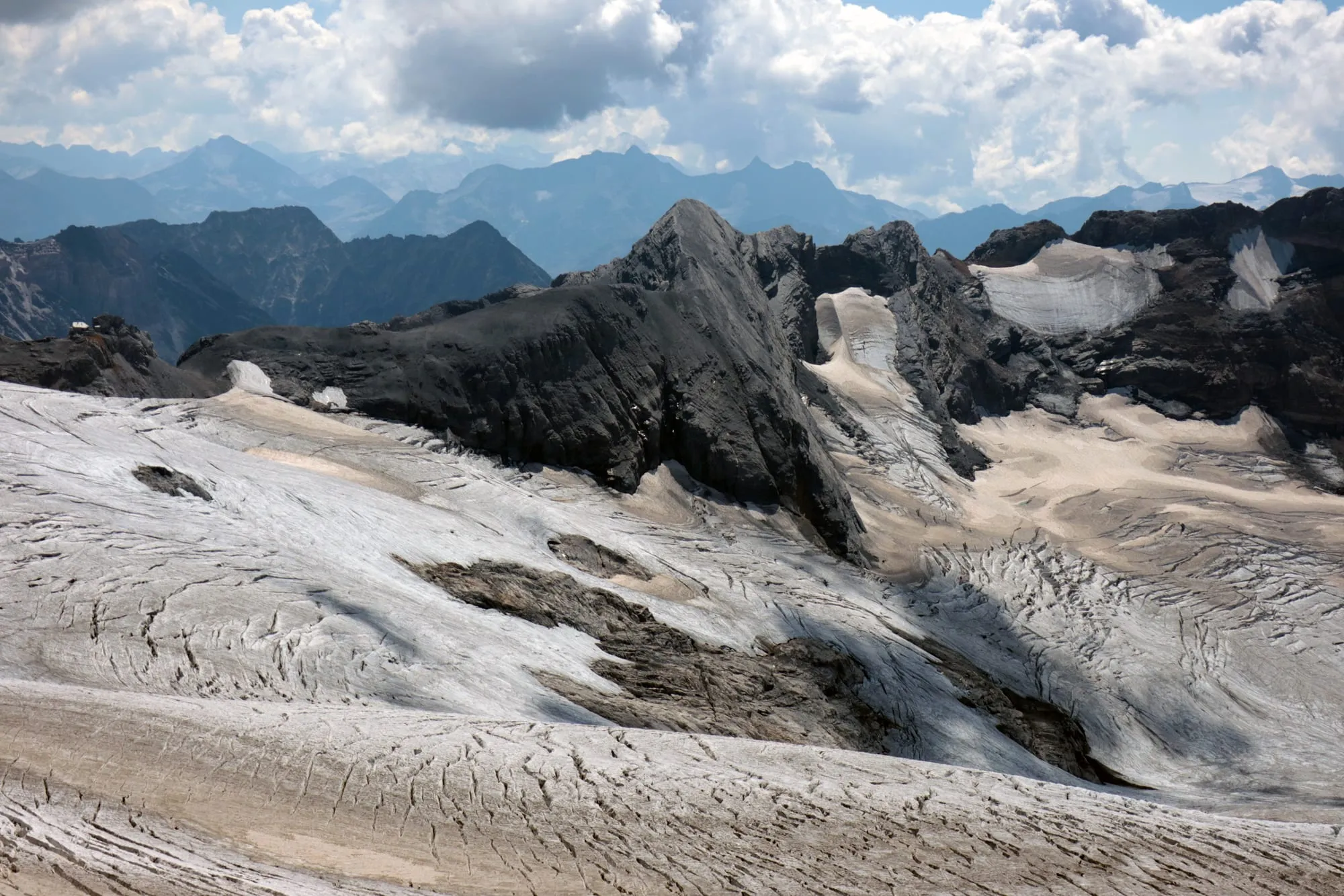 A wide view across the glacier plateau to black rock ridges and the ranges of the central Alps beyond.