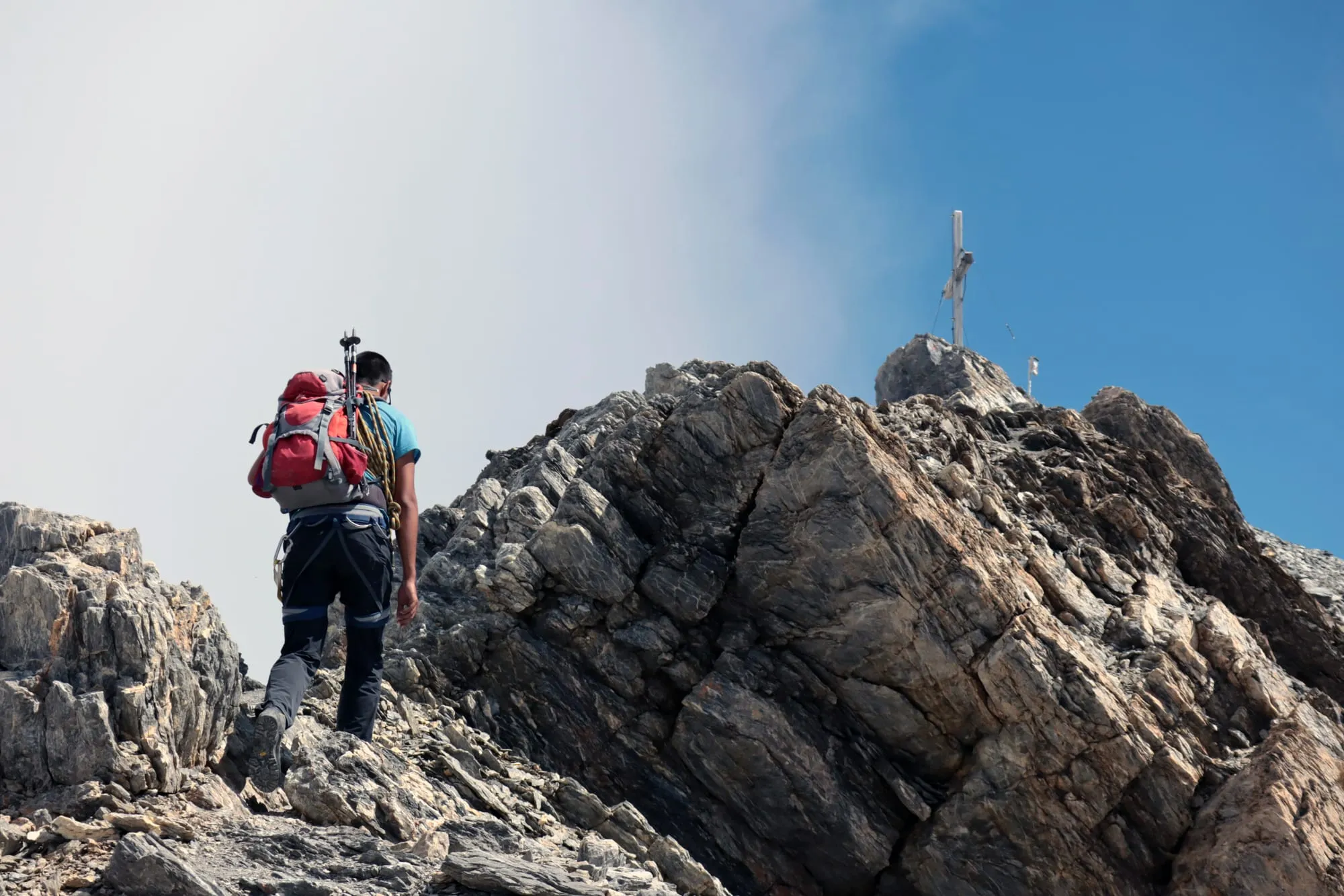 A mountaineer with a red pack climbs the last rocky ridge towards the Clariden summit cross.