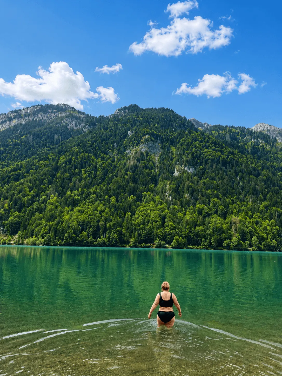 Wading waist-deep into the clear green lake, forested slopes and blue sky filling the far shore.