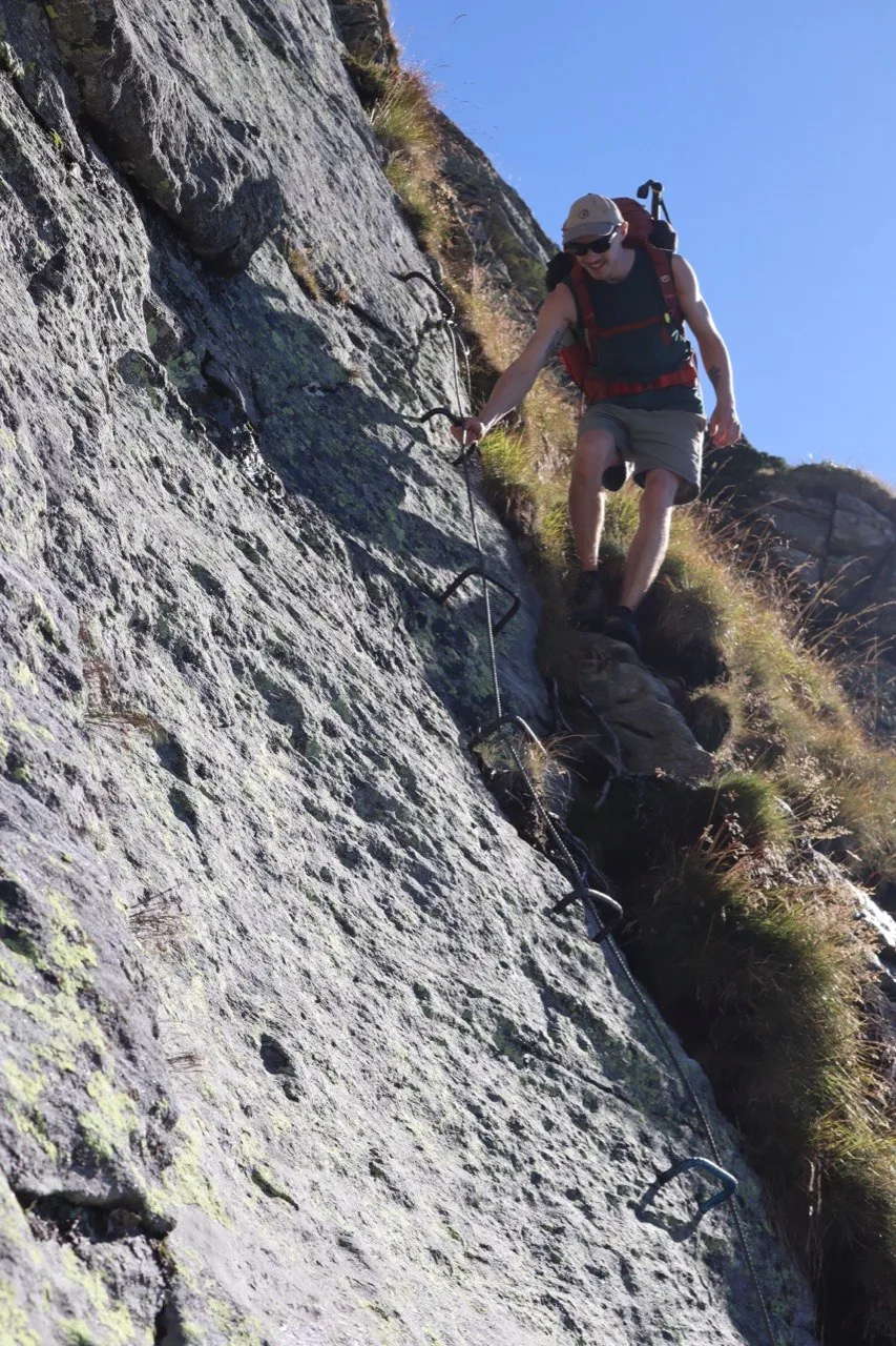 Hiker downclimbing a steep rock face on metal rungs and a fixed chain.