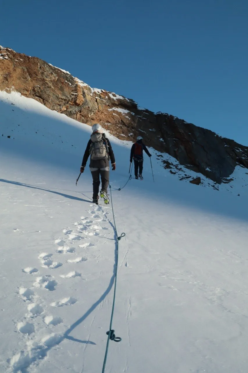 Two roped climbers crossing a snowfield toward a rock band under a blue sky.