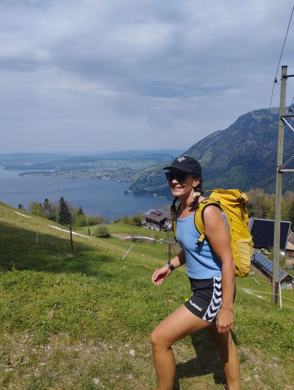 A smiling hiker with a yellow backpack, sunglasses, and a cap stands in a meadow high above the lake.