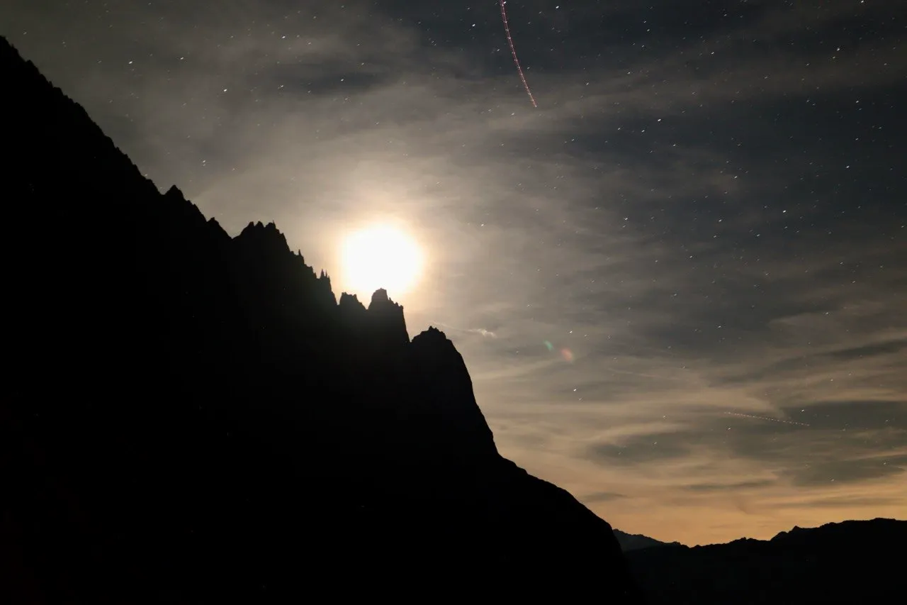 The moon glows behind a jagged mountain silhouette under a starry sky.