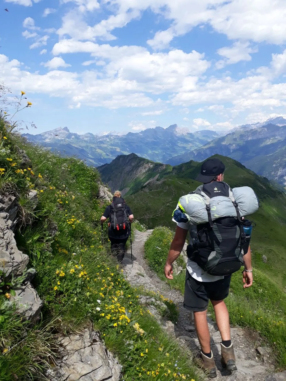 Two hikers follow a narrow path lined with yellow alpine flowers along a ridge with a sweeping view into the mountains.