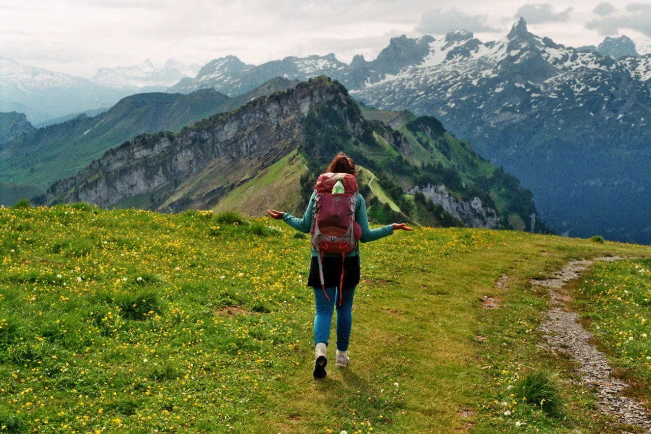 A hiker with a red backpack spreads her arms on a flower-strewn alpine meadow, facing the snow-dusted spikes of the Mythen range.