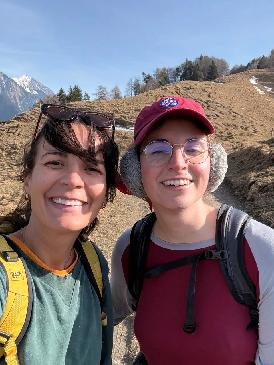 Two smiling hikers take a selfie on a sunny grassy slope, snowy peaks behind them.