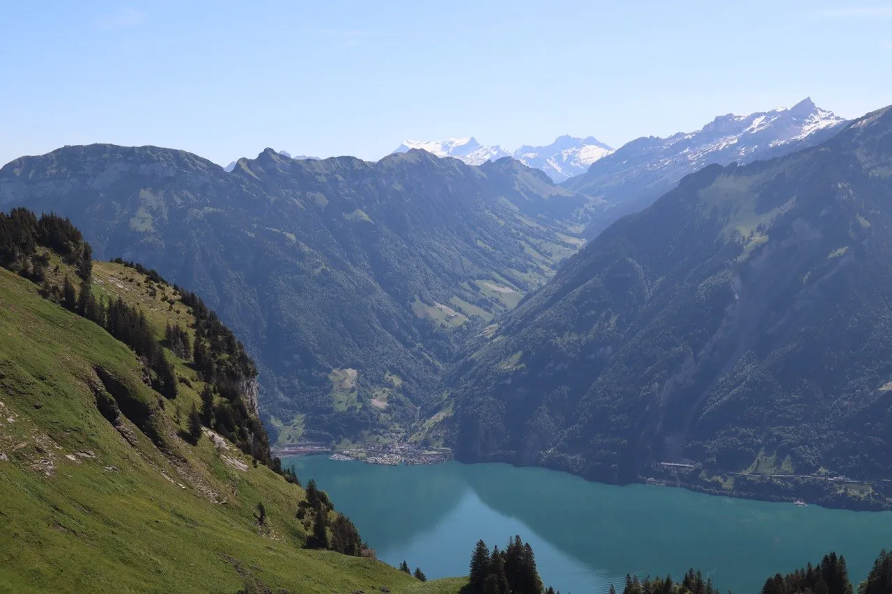 A green valley opens onto a turquoise lake, with snow-capped peaks lining the horizon.