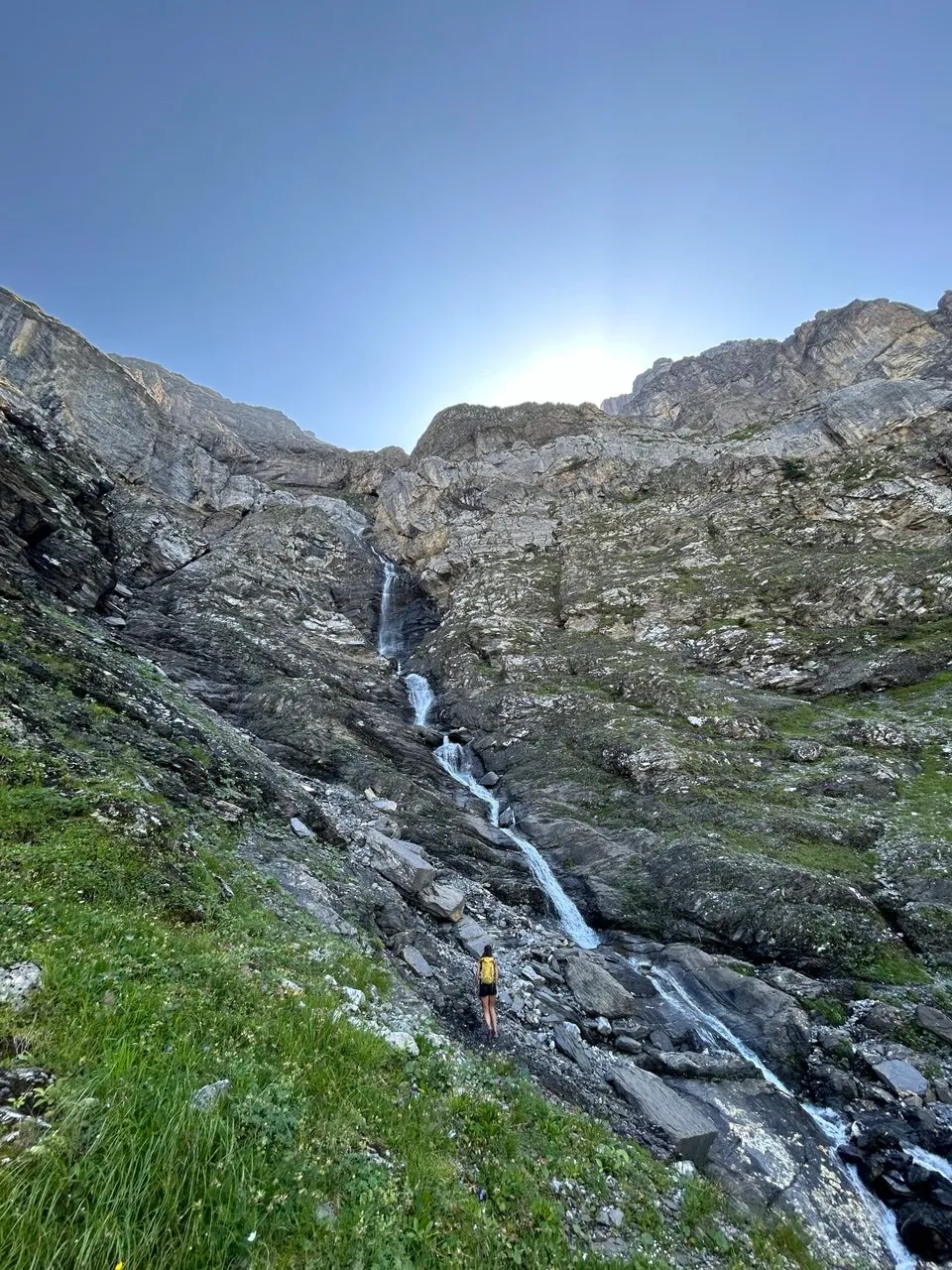 A tiny figure stands before a tall, multi-tiered waterfall cascading down a rock face.