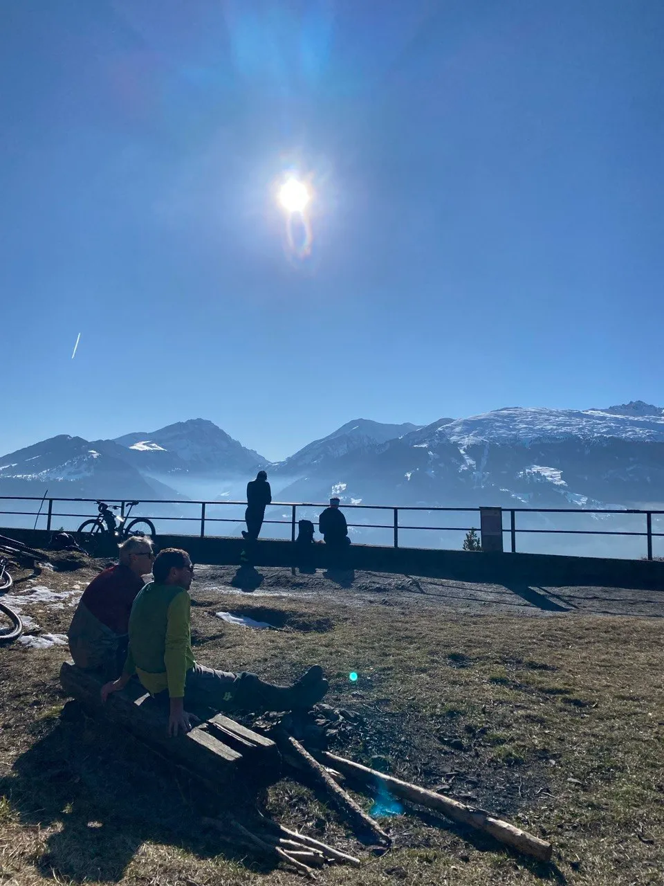 A group rests on wooden logs at the summit, looking over the fence at snow-capped mountains in the sun.