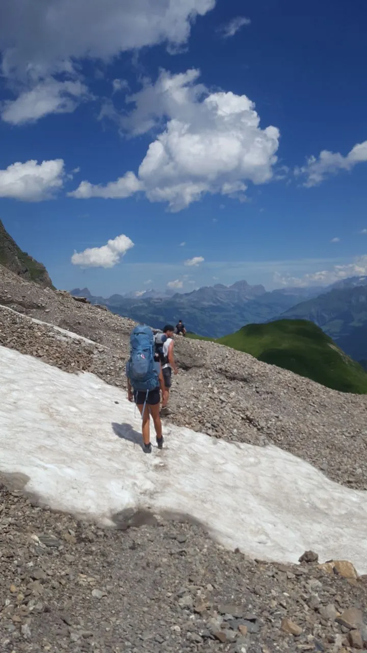 Hikers cross a summer snowfield over scree near the pass, with hazy mountain ranges in the distance.