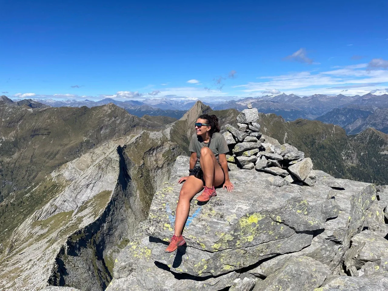 Hiker in sunglasses sitting on a rocky summit next to a cairn, mountains all around.
