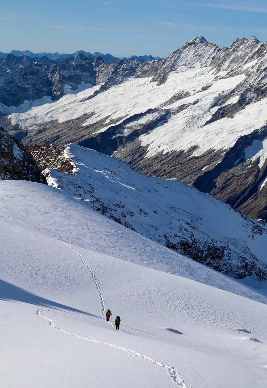 Two tiny figures on a vast glacier slope before a sea of peaks.
