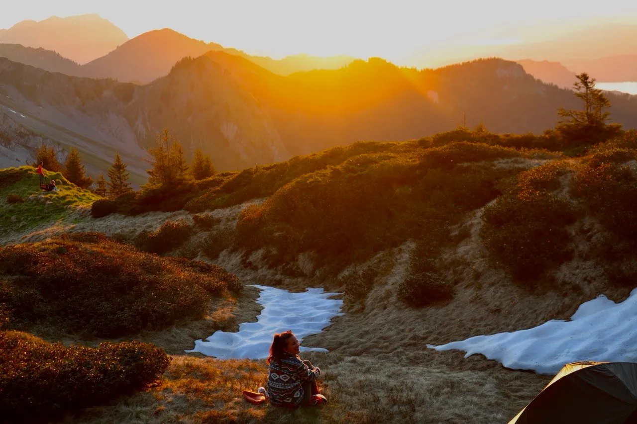 Una persona sentada en una ladera de hierba contempla la puesta de sol sobre siluetas de montañas escalonadas, con una mancha de nieve debajo.