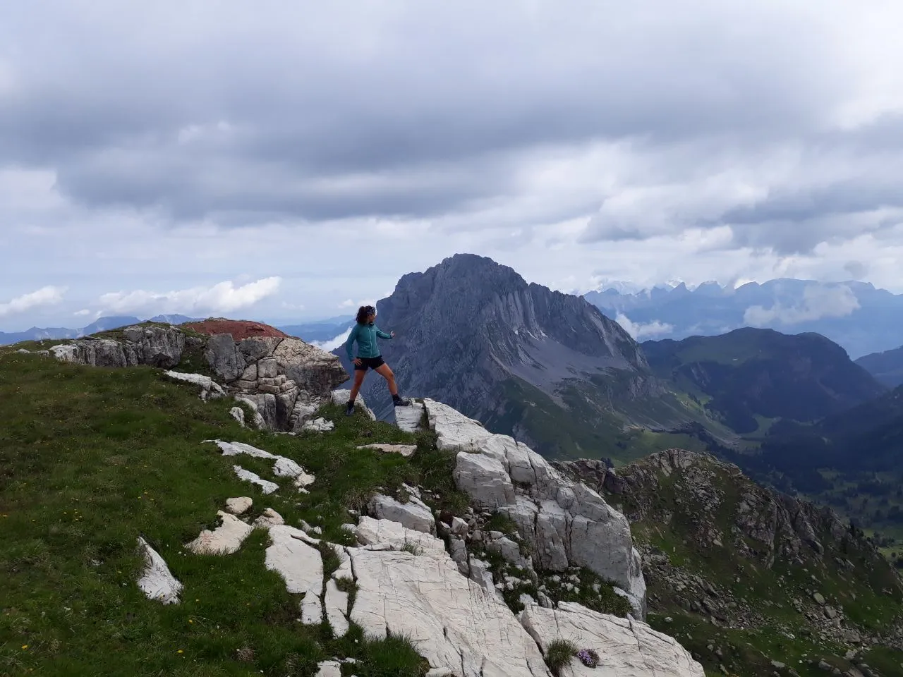 A hiker in a teal jacket stepping across a pale limestone ridge, a steep rocky peak and hazy distant ranges rising behind under a cloudy sky.