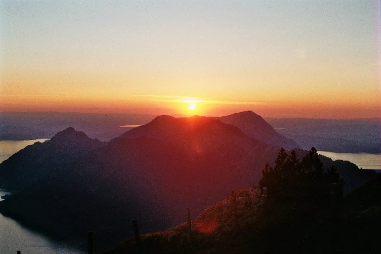 Sunset over layered, dark-silhouetted peaks, the sun sinking into an orange-red sky above the lake.