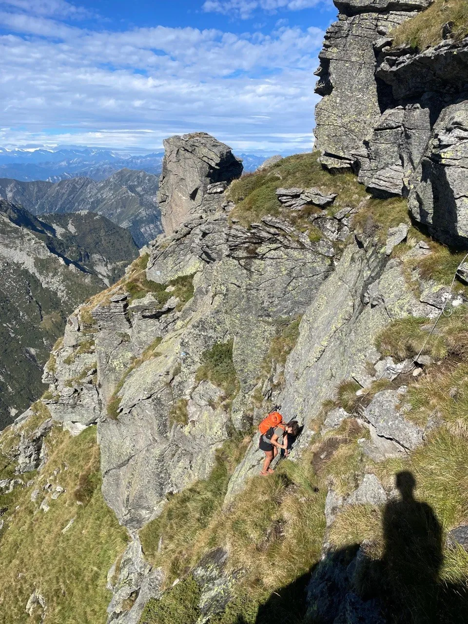 Two hikers scrambling up a steep grassy flank below craggy rock towers.