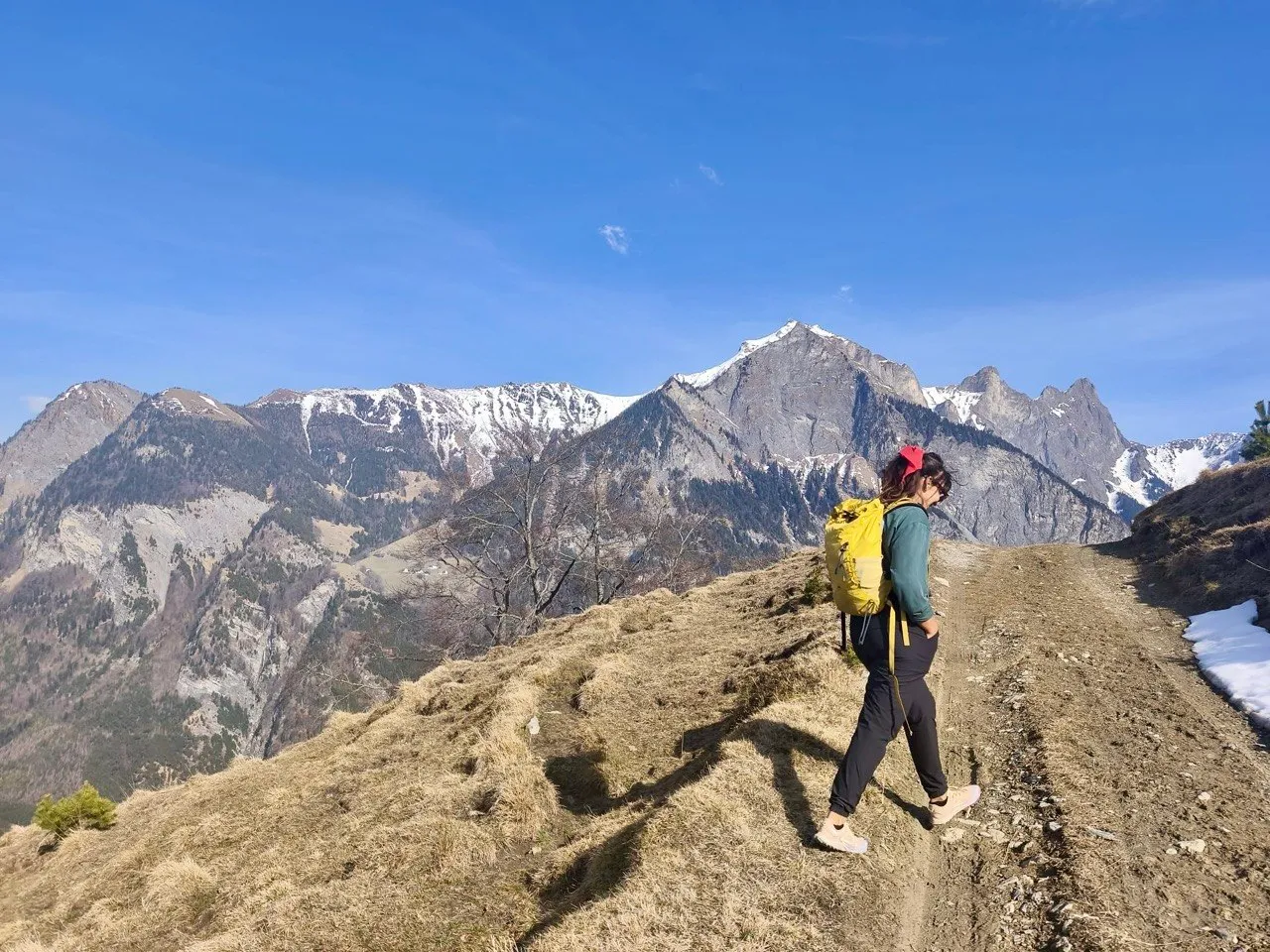 A hiker with a yellow backpack walks a grassy ridge path toward snow-streaked rocky peaks under a clear blue sky.