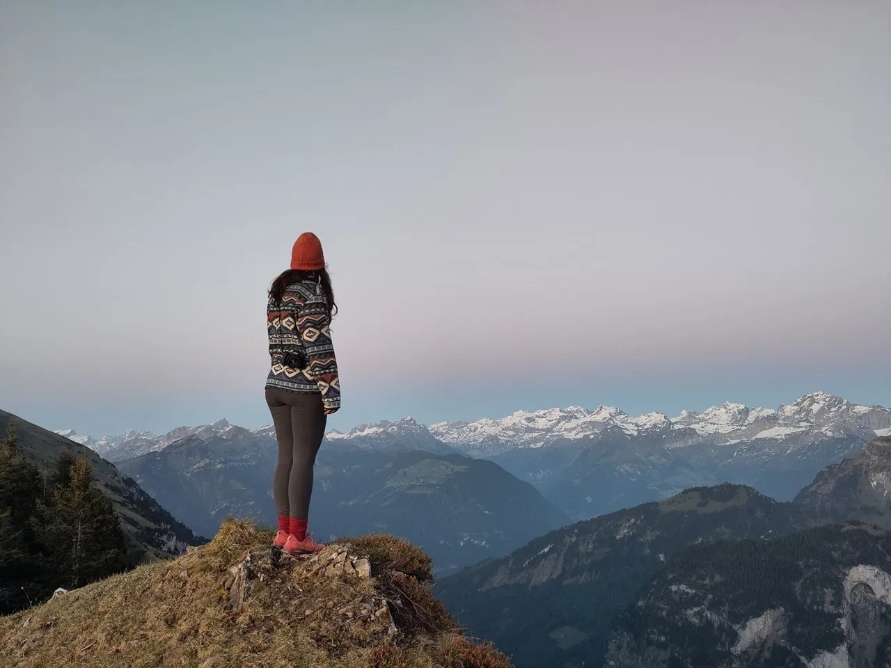 A hiker in a red beanie stands on a rocky outcrop at dusk, camera in hand, facing a horizon of snow-capped peaks.