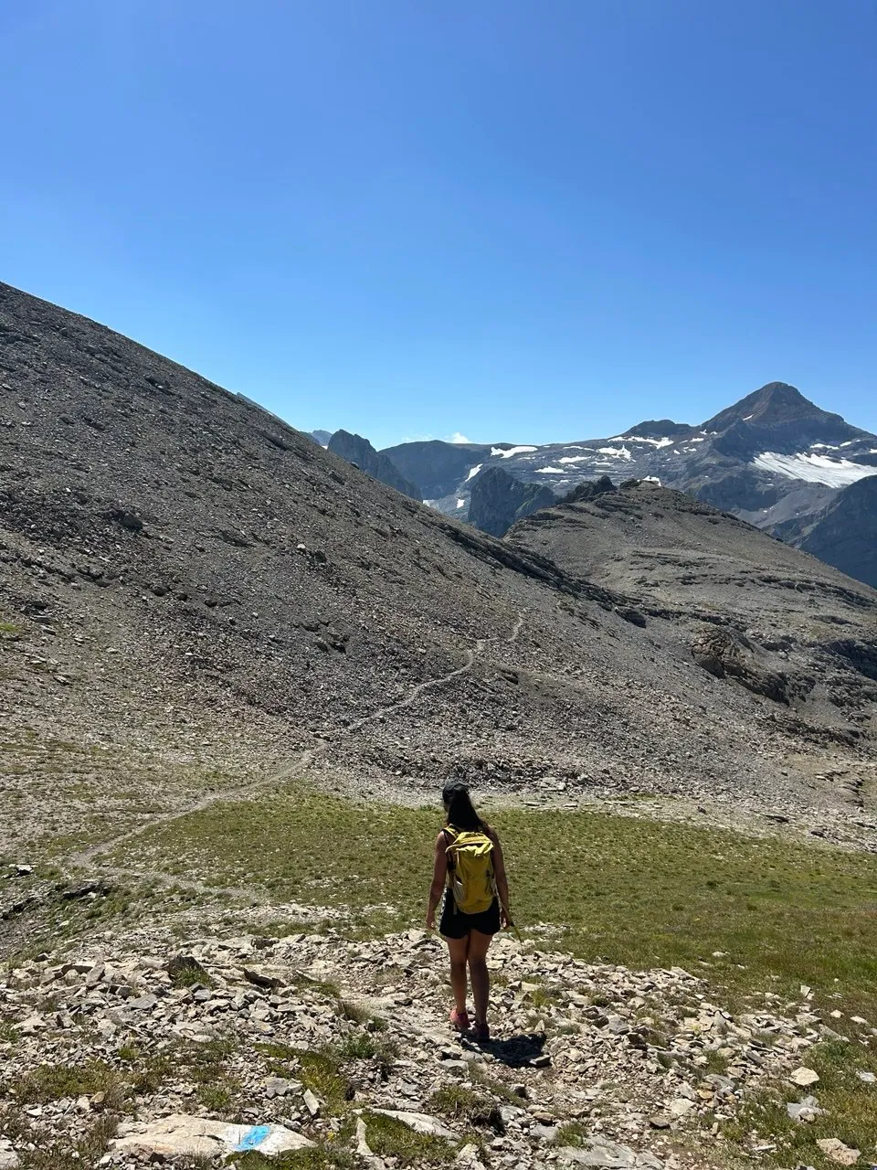 A hiker with a yellow backpack walks a path through a wide scree field, surrounded by rugged mountain ranges.