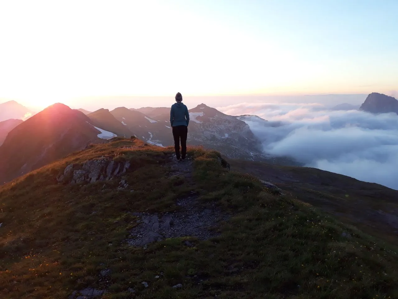 A hiker standing on a grassy ridge at dawn, gazing over a sea of cloud filling the valley with the sun rising low over the peaks.