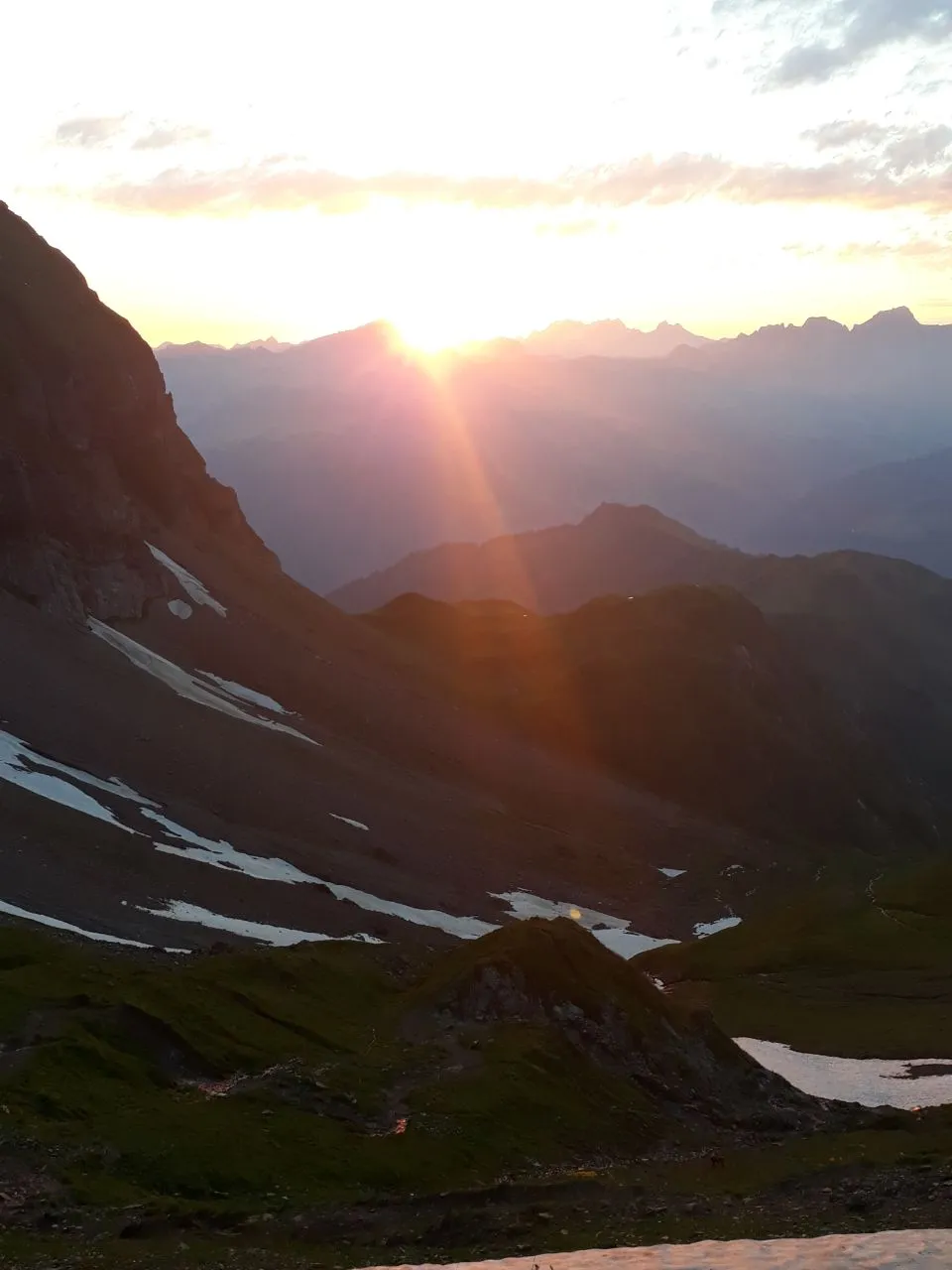 The sun sits low over layered mountain ranges, bathing a high valley with a small mountain lake in warm light.