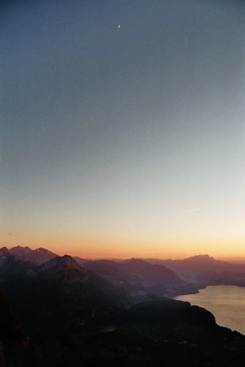 Dusk over the lake and mountain ranges, a thin crescent moon above the last orange band on the horizon.