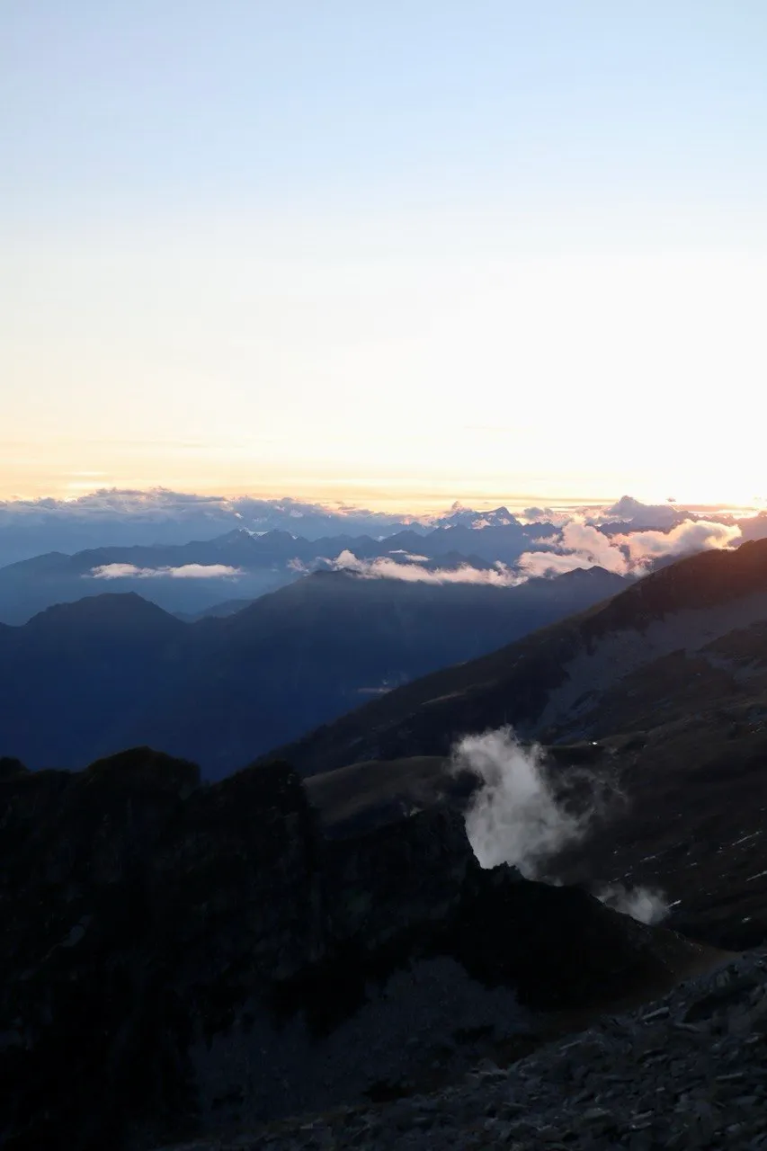 Sunrise over layered, misty mountain ridges with a small cloud rising from the slope.